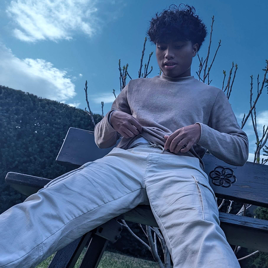 Portrait von broizznico auf einer Parkbank. Im Hintergrund sind Hecken und ein blauer Himmel mit wenigen Wolken.