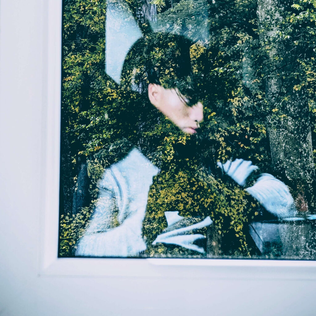 Ein Selfie vom Musiker broizznico in der Reflexion einer Fensterscheibe. Zum Teil ist im Foto, das von innen nach außen fotografiert wurde, die Außenwelt vor dem Fenster sichtbar: Bäume mit grünen Blättern in einem warmen Licht.
